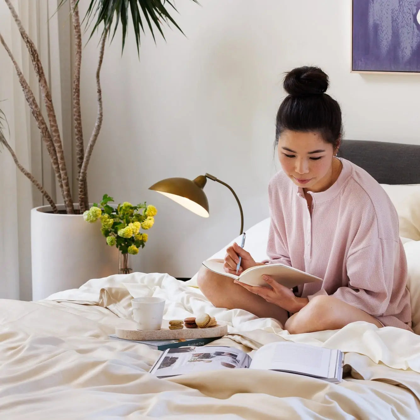 A woman sits on a bed in a bright, modern bedroom, writing in a notebook. She is wearing a light pink long-sleeve top.