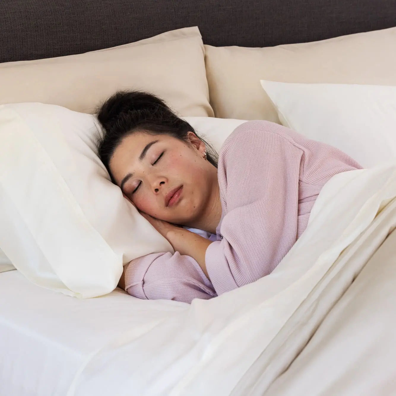 A woman sleeping soundly on a bed with white pillows and duvet, wearing a light pink ribbed long-sleeve top. 