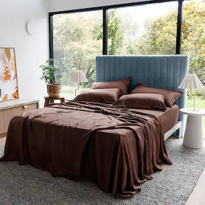 A bed dressed in dark brown bedding and matching pillows, positioned in a modern bedroom with a blue upholstered headboard, large windows showing green trees outside, and decorative side tables and plants. 