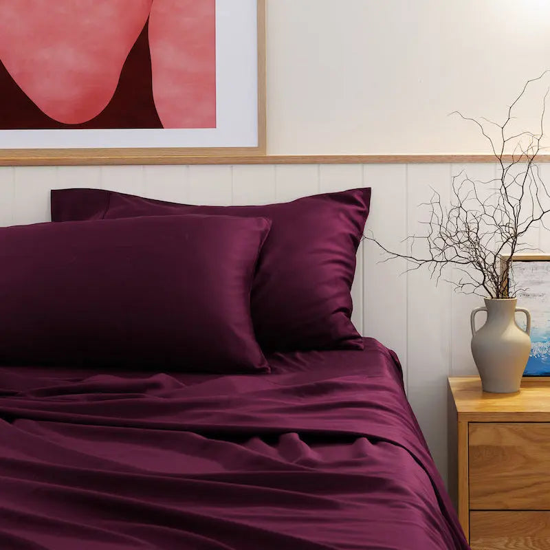 A bed dressed in deep burgundy satin sheets and matching pillowcases, styled in a bedroom with a light wood headboard, a framed abstract pink artwork on the wall, and a small wooden nightstand with a vase of branches. 