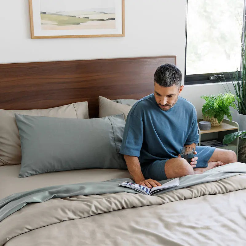 A man in a blue t-shirt and shorts sits on a bed with gray and beige bedding, reading a book in a bright, modern bedroom with plants and a wooden headboard. 