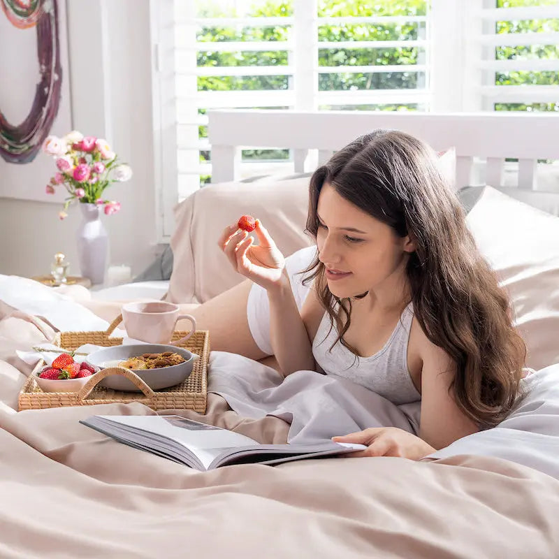 A woman with long brown hair lies on a bed reading a book, holding a strawberry. A breakfast tray with a cup, cereal, and more strawberries is placed on the bed beside her. 