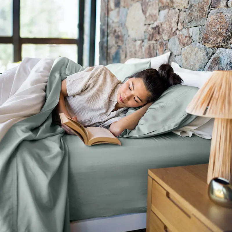 Woman reading a book in bed with a stone wall and window in the background. 