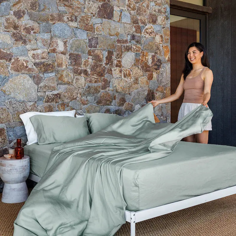 Woman arranging green bedding in a bedroom with a stone wall. 