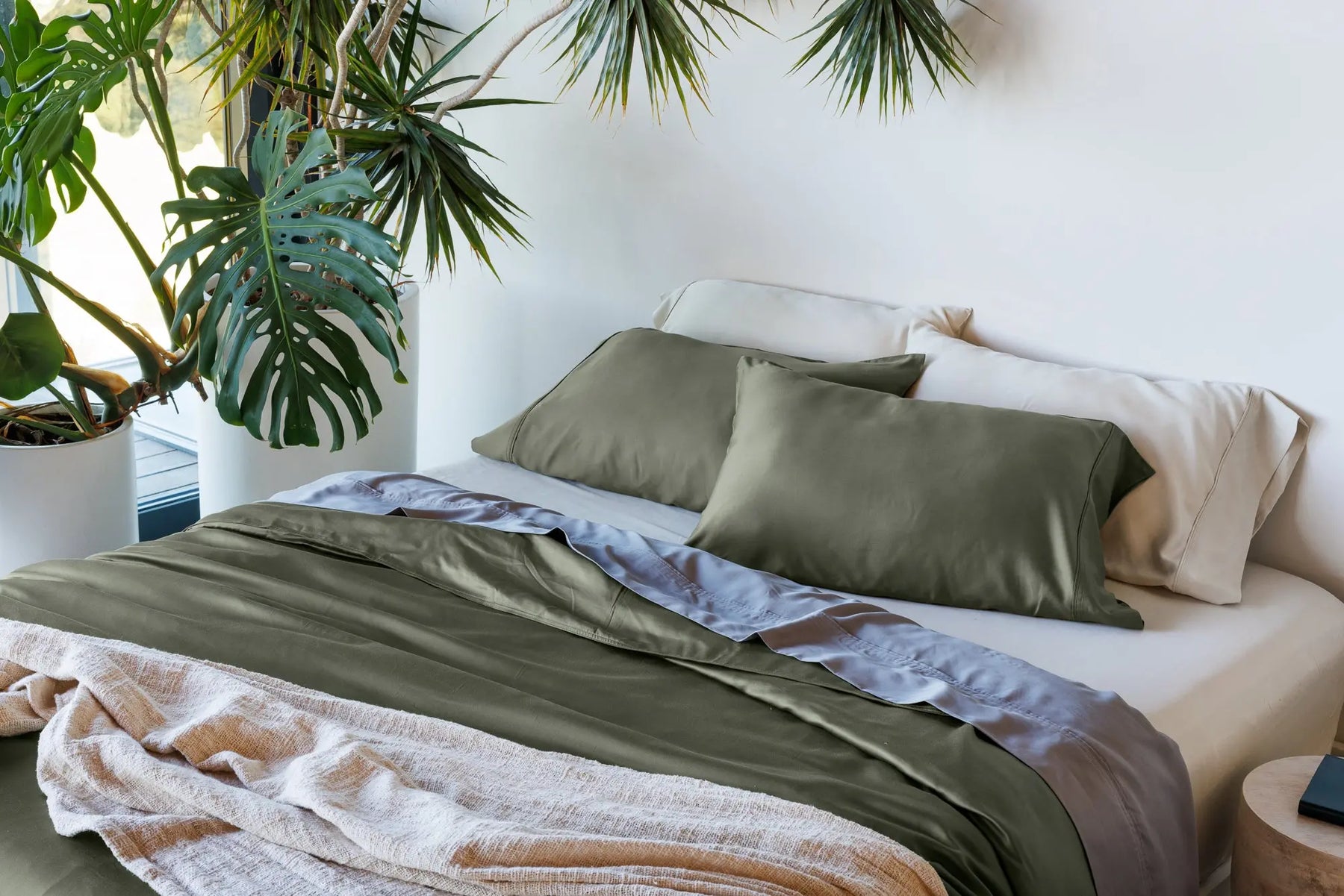 A neatly made bed in a bright bedroom setting, styled with olive green bedding, cream pillows, a light blue duvet layer, and a beige textured throw blanket. Large potted plants are visible on the left, and the background is a clean white wall. 