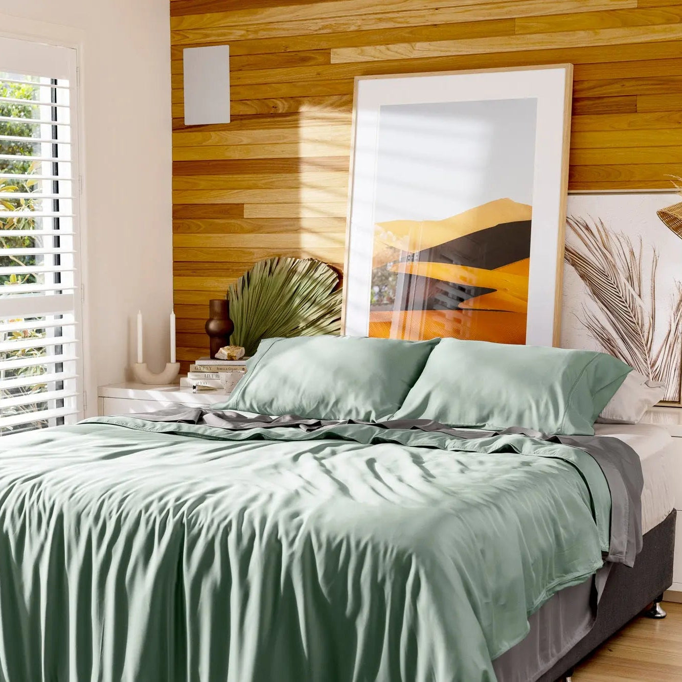 A bed dressed in a sage green bedding set with matching pillows, styled in a modern bedroom with a wood-paneled wall, white shutters, and decorative wall art. 