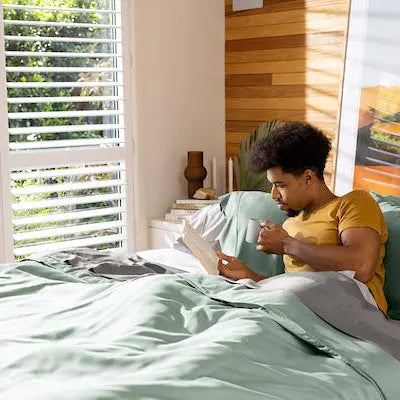 Person reading a book on a bed with green bedding in a bright room. 