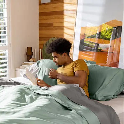 Person reading a book on a bed with green bedding in a bright room. 