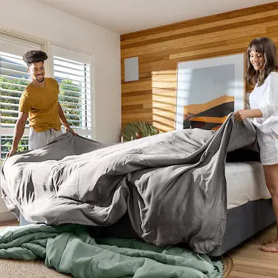 Two people setting up a bed in a bright bedroom with wooden walls and large windows. 