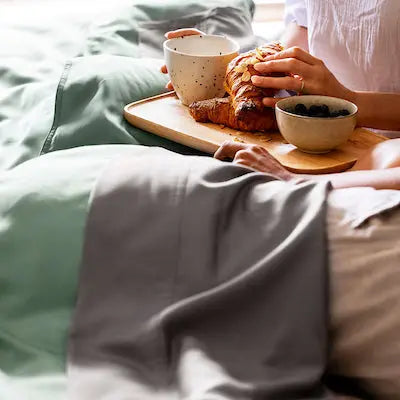 Person sitting on a bed with a breakfast tray, holding a croissant and a cup. 