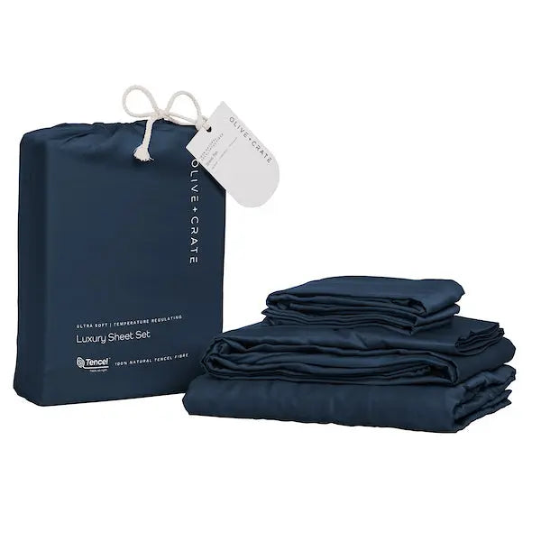 A navy blue luxury sheet set displayed next to its branded drawstring storage bag. The set includes neatly folded sheets and pillowcases, and the bag features a white hanging tag with the brand name Olive and Crate. French Navy sheet set 