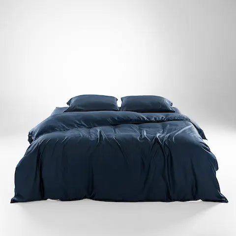 A neatly made bed with dark navy blue bedding, including a duvet cover and two matching pillows, photographed in a clean studio setting with a light gray gradient background. 