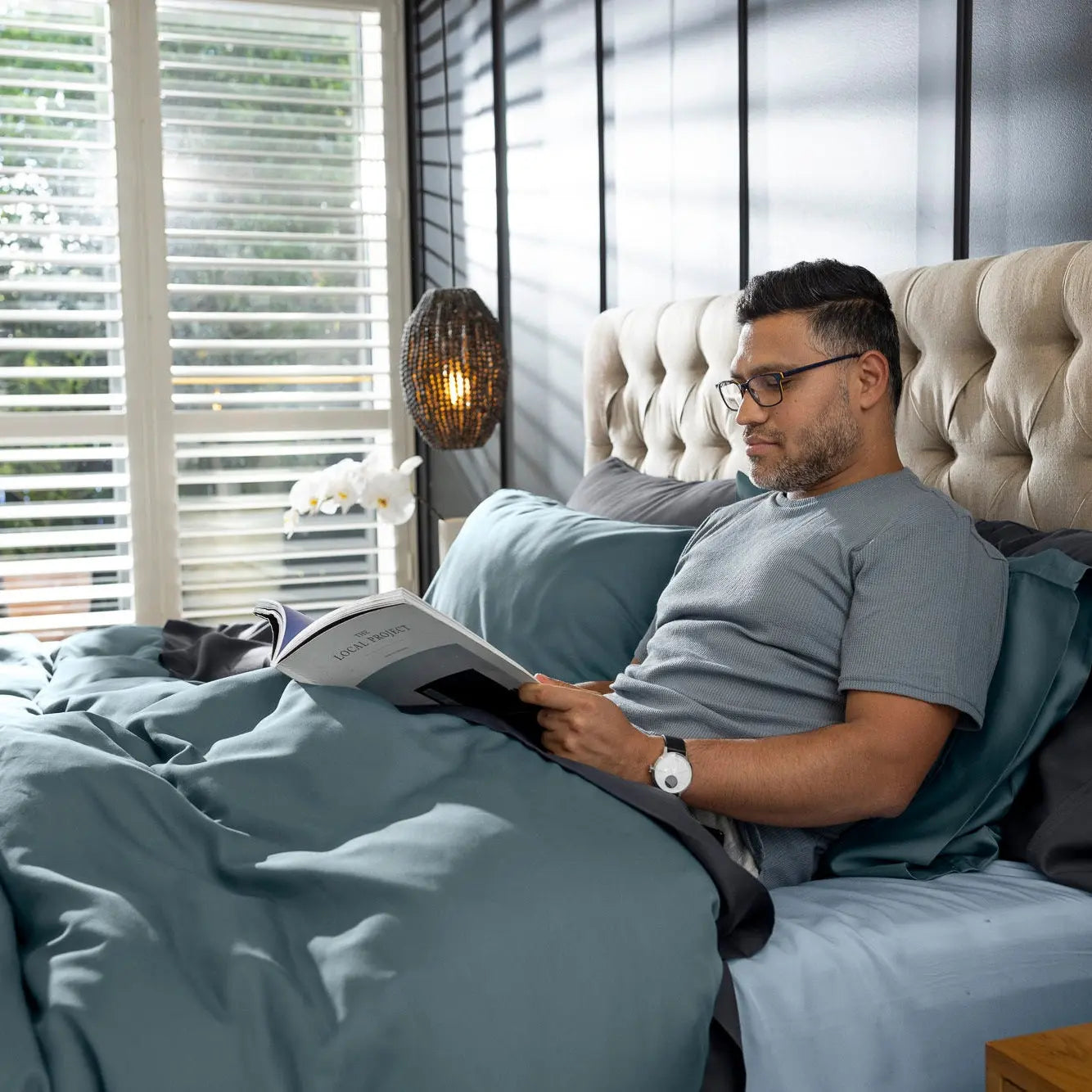 A man wearing glasses lies in bed reading a magazine, surrounded by slate blue bedding, a tufted beige headboard, and a modern bedroom setting. 