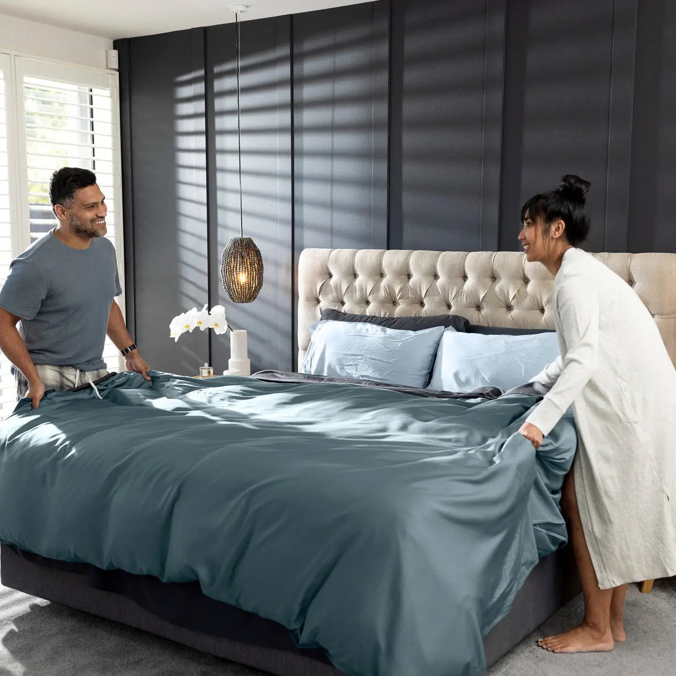 A man and a woman in a modern bedroom setting, adjusting a slate blue duvet cover on a bed with a tufted headboard. The room features dark paneled walls, hanging pendant lights, a wooden nightstand, and a window with blinds. 